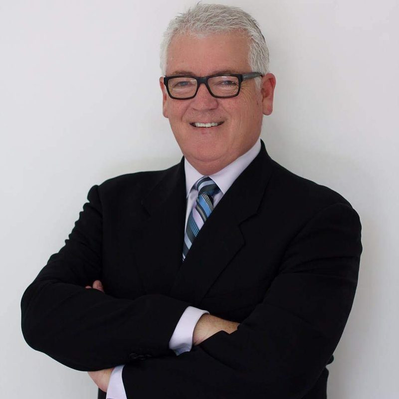 Michael Corridan, in a dark suit with a striped blue tie and black-framed glasses, arms folded, against a plain white background