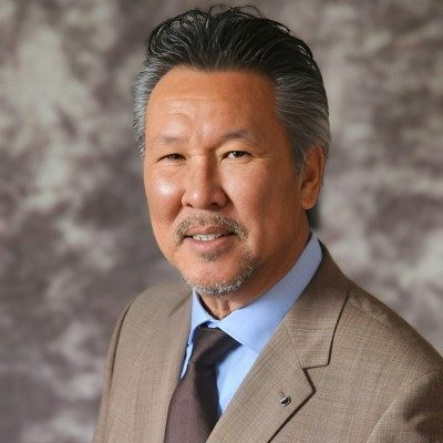 Steve Lim, in a tan jacket over a blue shirt and dark tie, against a mottled brown studio background