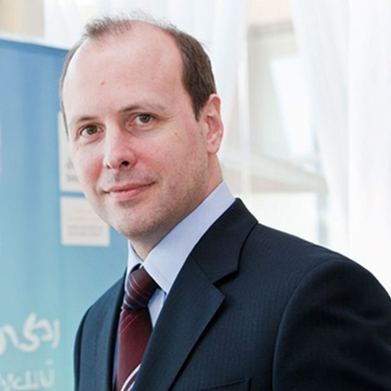Dr Nigel Rea, in a navy suit and dark red tie, photographed at an event beside a blue banner printed in Arabic